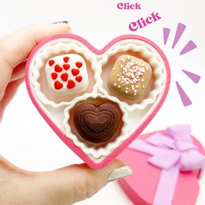 May include: A pink heart-shaped box filled with three decorative chocolates. One chocolate is white with red hearts, another is tan with pink sprinkles, and the last is a brown heart shape. The box is held in a hand.