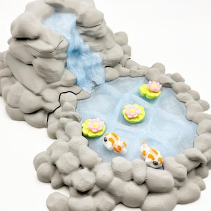 Puede incluir: Una escena de jard&iacute;n zen en miniatura con una cascada que desemboca en un estanque. El estanque tiene nen&uacute;fares con flores rosas y amarillas, y dos carpas koi naranjas y blancas. Las rocas circundantes son grises.