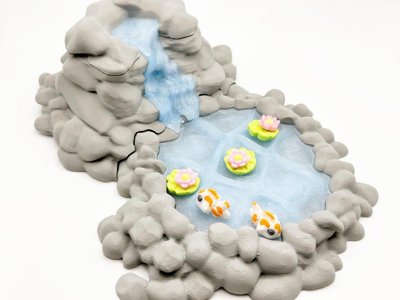 Puede incluir: Una escena de jard&iacute;n zen en miniatura con una cascada que desemboca en un estanque. El estanque tiene nen&uacute;fares con flores rosas y amarillas, y dos carpas koi naranjas y blancas. Las rocas circundantes son grises.
