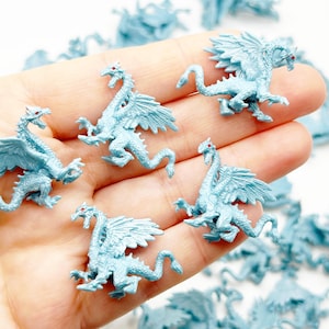 Peut inclure: Une main tenant une collection de petites figurines de dragons en plastique bleu clair. Les dragons ont des ailes, des queues et des yeux rouges.