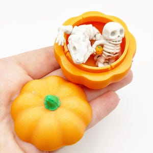 Peut inclure: Un récipient orange en forme de citrouille avec une tige verte, contenant un squelette blanc et un squelette de dinosaure. Le couvercle est à côté du récipient. Décor sur le thème d'Halloween.