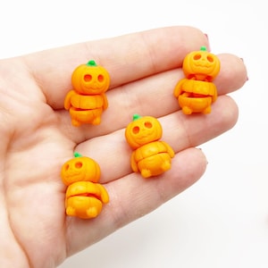 以下が含まれることがあります： 緑色の茎が付いたオレンジ色のカボチャの形をした小さな置物。ミニチュアのかぼちゃは彫刻された顔とセグメント化された体を持っています。置物は明るい背景に表示されており、ハロウィーンまたは秋のテーマを示唆しています。