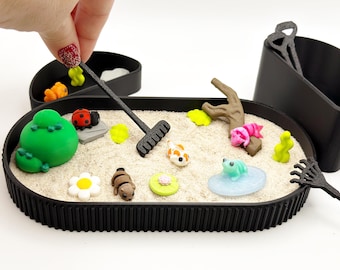 Op de afbeelding: Een ovaal zwart dienblad gevuld met lichtgekleurd zand, met miniatuurfiguurtjes waaronder een groene heuvel, een lieveheersbeestje, een witte bloem, een bruin dier, een waterlelieblad, een koikarper, een kikker op een blauwe schijf en een bruine tak, met twee zwarte harken.