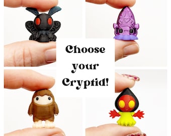 May include: A collage of four miniature cryptid figurines: a black moth-like creature, a purple squid-like figure, a brown yeti, and a red and yellow humanoid. The text "Choose your Cryptid!" is in the center.