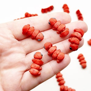 May include: A hand holding several small, red, segmented, plastic toy insects. The insects have black eyes and are arranged in a cluster on the palm of the hand.