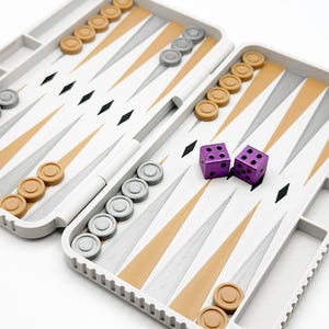May include: A white and tan backgammon set with gray and tan game pieces and two purple dice. The board is open, revealing the playing surface with a geometric design. The set is designed for travel.