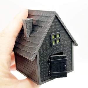 Peut inclure: Une petite maison mod&egrave;le imprim&eacute;e en 3D de couleur noire, dot&eacute;e d'un toit textur&eacute;, d'une chemin&eacute;e, d'une fen&ecirc;tre unique avec un int&eacute;rieur vert clair et d'une porte &agrave; charni&egrave;res entrouverte. La structure pr&eacute;sente des d&eacute;tails de bardage horizontal.