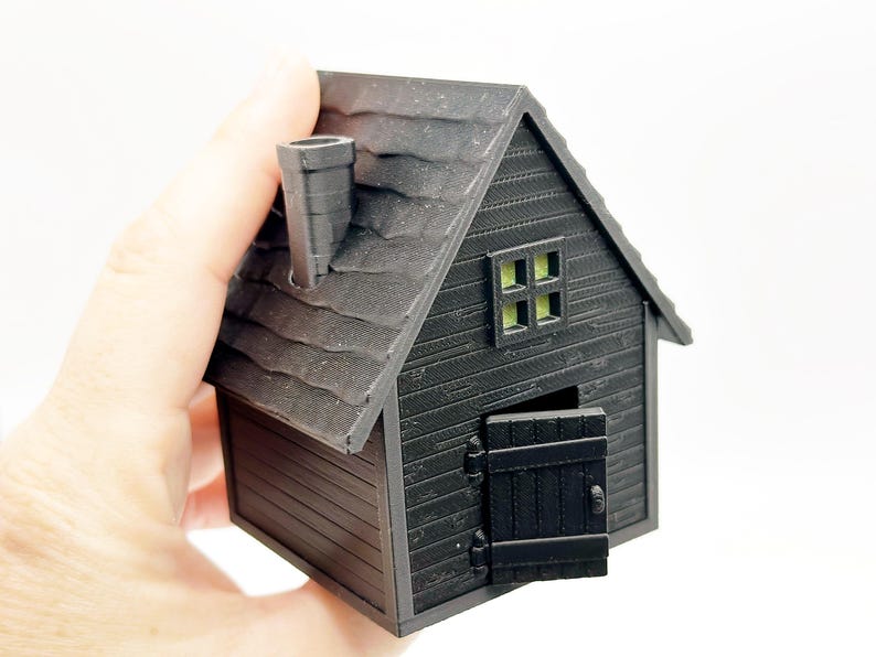 Peut inclure: Une petite maison mod&egrave;le imprim&eacute;e en 3D de couleur noire, dot&eacute;e d'un toit textur&eacute;, d'une chemin&eacute;e, d'une fen&ecirc;tre unique avec un int&eacute;rieur vert clair et d'une porte &agrave; charni&egrave;res entrouverte. La structure pr&eacute;sente des d&eacute;tails de bardage horizontal.
