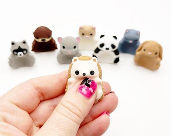 Op de afbeelding: Een verzameling kleine, gegoten dierenfiguurtjes waaronder een witte egel, een grijze wasbeer, een bruine otter, een grijze chinchilla, een beige hamster, een zwart-witte panda, een blauwe kat en een beige konijn met gladde, matte oppervlakken.