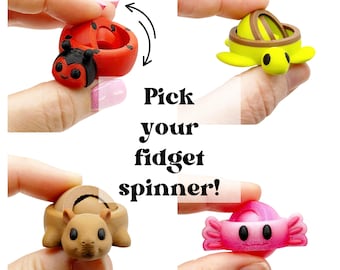 Puede incluir: Cuatro pequeños fidget spinners de plástico con formas de animales y dulces, incluyendo una mariquita roja, una tortuga verde, una ardilla beige y un caramelo rosa. El texto "Pick your fidget spinner!" está centrado con "spin spin" y una flecha.