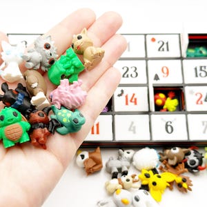 May include: A collection of small, colourful plastic creature figurines in various shapes and colours, including green, pink, black, and yellow, alongside a black and white advent calendar box with numbered compartments containing additional figures.