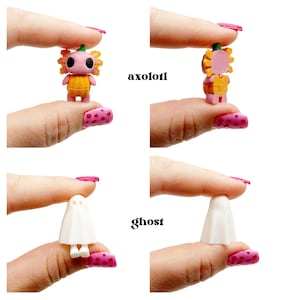 Peut inclure: Une figurine miniature d'axolotl rose portant un costume de citrouille orange et une petite figurine de fant&ocirc;me blanc. L'axolotl a de grands yeux noirs et des branchies jaunes. Le fant&ocirc;me est blanc avec deux petits trous pour les yeux.