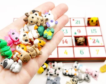 Op de afbeelding: Een verzameling kleine, kleurrijke gummen in dierenvorm, waaronder een geit, eenhoorn, axolotl, tijger en wasbeer. Deze miniatuurfiguurtjes worden in een handpalm gehouden naast een genummerde adventskalender met open vakjes.