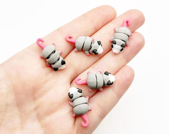 Op de afbeelding: Vijf kleine, gearticuleerde opossum-figuurtjes met grijze lichamen, witte gezichten, zwarte oogvlekken en roze staarten. Elk miniatuurdier heeft een gesegmenteerd ontwerp dat beweging mogelijk maakt, met gedetailleerde texturen op de rug.