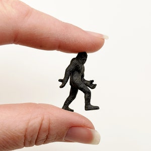 May include: A small black plastic figurine of Bigfoot walking, held between two fingers.