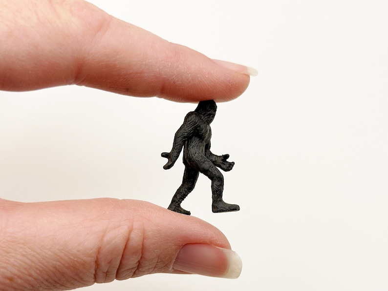 May include: A small black plastic figurine of Bigfoot walking, held between two fingers.