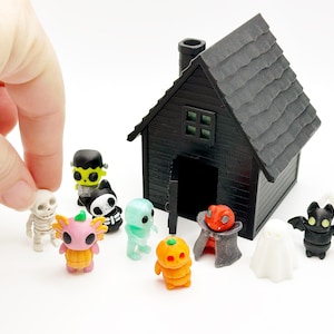 Peut inclure: Une maison miniature noire avec une chemin&eacute;e et une porte ouverte, entour&eacute;e de petites figurines en plastique incluant un squelette, un monstre vert, un chat noir et blanc, un fant&ocirc;me bleu clair, un axolotl rose dans une citrouille, une citrouille orange, un vampire, un fant&ocirc;me blanc et une chauve-souris noire.