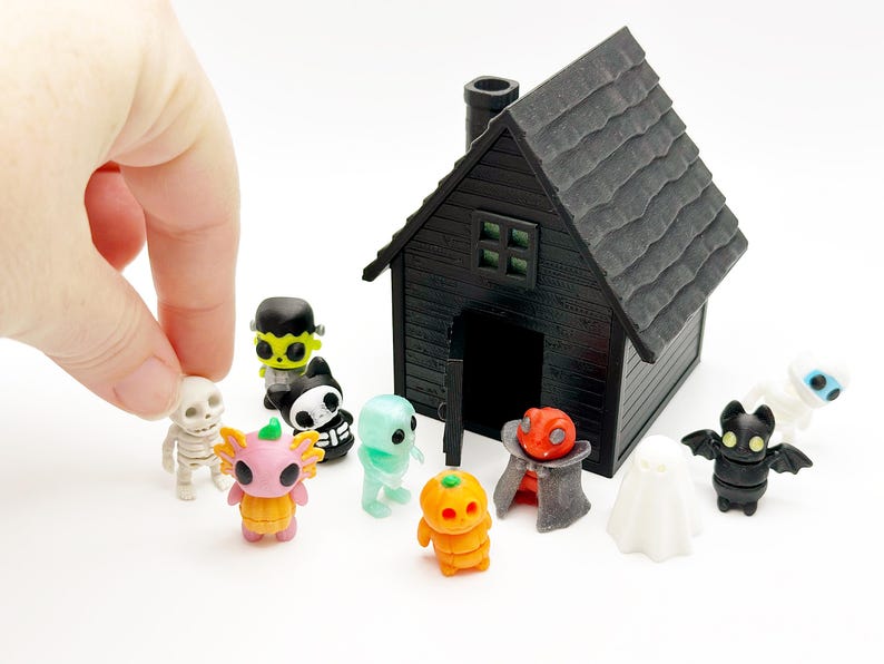 Peut inclure: Une maison miniature noire avec une chemin&eacute;e et une porte ouverte, entour&eacute;e de petites figurines en plastique incluant un squelette, un monstre vert, un chat noir et blanc, un fant&ocirc;me bleu clair, un axolotl rose dans une citrouille, une citrouille orange, un vampire, un fant&ocirc;me blanc et une chauve-souris noire.