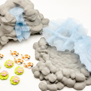 Puede incluir: Una escena de estanque en miniatura con formaciones rocosas grises, una cascada azul y varias peque&ntilde;as figuras de peces koi con marcas naranjas y blancas. Tambi&eacute;n se incluyen nen&uacute;fares verdes con flores rosas y amarillas.