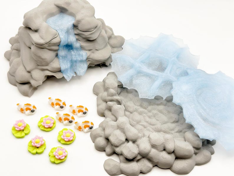 Puede incluir: Una escena de estanque en miniatura con formaciones rocosas grises, una cascada azul y varias peque&ntilde;as figuras de peces koi con marcas naranjas y blancas. Tambi&eacute;n se incluyen nen&uacute;fares verdes con flores rosas y amarillas.