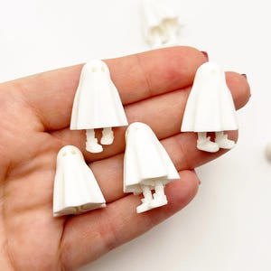 May include: A collection of small, white ghost figurines. Each ghost has a simple, rounded body and tiny feet. The ghosts are arranged in a hand, suggesting a craft or decorative item, perfect for Halloween or spooky-themed projects.