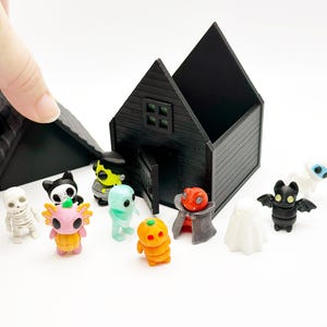Peut inclure: Une maison miniature noire avec un toit amovible et une collection de petites figurines color&eacute;es, dont un squelette, un axolotl rose, un monstre vert, une citrouille, un vampire, un fant&ocirc;me, une chauve-souris et une momie sur fond blanc.