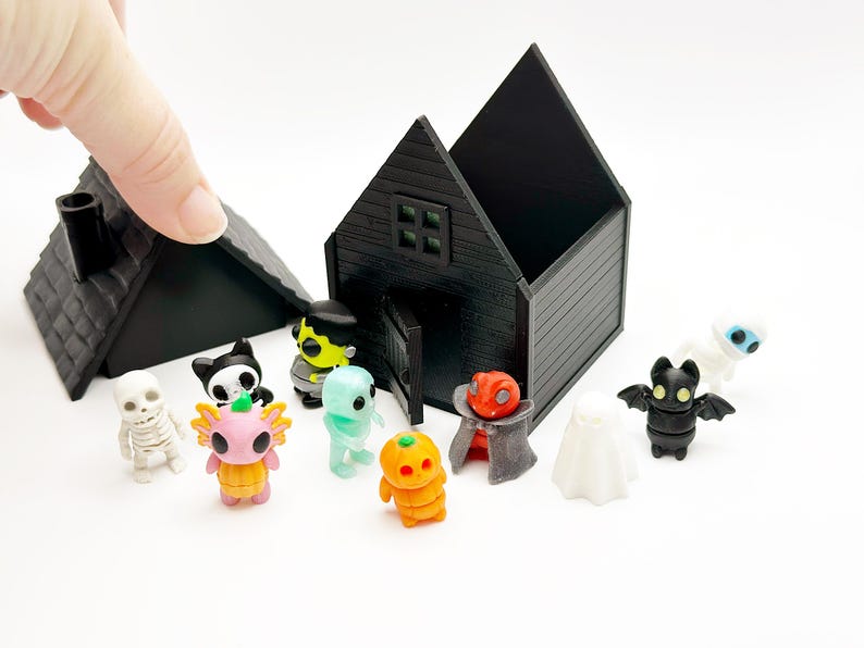 Peut inclure: Une maison miniature noire avec un toit amovible et une collection de petites figurines color&eacute;es, dont un squelette, un axolotl rose, un monstre vert, une citrouille, un vampire, un fant&ocirc;me, une chauve-souris et une momie sur fond blanc.