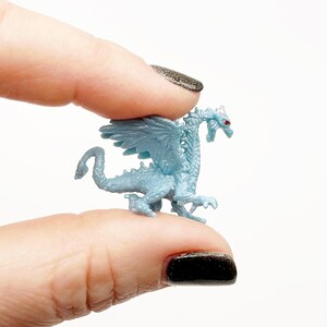Peut inclure: Une petite figurine de dragon en plastique bleu clair avec des yeux rouges. Le dragon a des ailes et est en position debout.