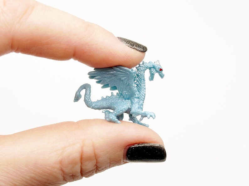 Peut inclure: Une petite figurine de dragon en plastique bleu clair avec des yeux rouges. Le dragon a des ailes et est en position debout.