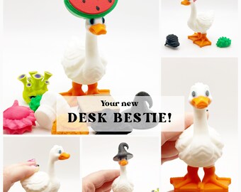 Op de afbeelding: Een wit ganzenbeeldje met oranje voeten en snavel, voorzien van verwisselbare accessoires waaronder een watermeloenhoed, een roze hanenkam en een zwarte heksenhoed. De tekst "Your new DESK BESTIE!" is over de afbeelding geplaatst.