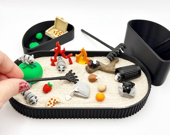 Op de afbeelding: Een zwarte ovale bak gevuld met lichtgekleurd zand met miniatuurfiguurtjes waaronder wasberen, een kampvuur, een pizzapunt, een aardbei, een visgraat, een boomstam en een zwarte prullenbak. Een zwarte hark is inbegrepen voor het zand.