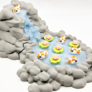 Puede incluir: Una escena de jard&iacute;n zen en miniatura con una cascada que desemboca en un estanque. El estanque est&aacute; poblado de peque&ntilde;os peces koi y nen&uacute;fares con flores rosas. Las rocas circundantes son grises.