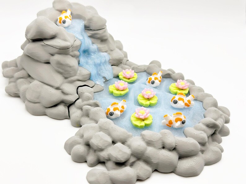 Puede incluir: Una escena de jard&iacute;n zen en miniatura con una cascada que desemboca en un estanque. El estanque est&aacute; poblado de peque&ntilde;os peces koi y nen&uacute;fares con flores rosas. Las rocas circundantes son grises.