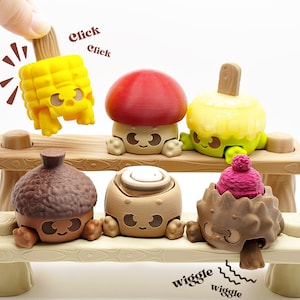 May include: A collection of whimsical, food-themed toys, including a corn on the cob, mushroom, and acorn, arranged on a miniature bench. The toys have smiling faces and are in various colors. The words "click" and "wiggle" are also visible.