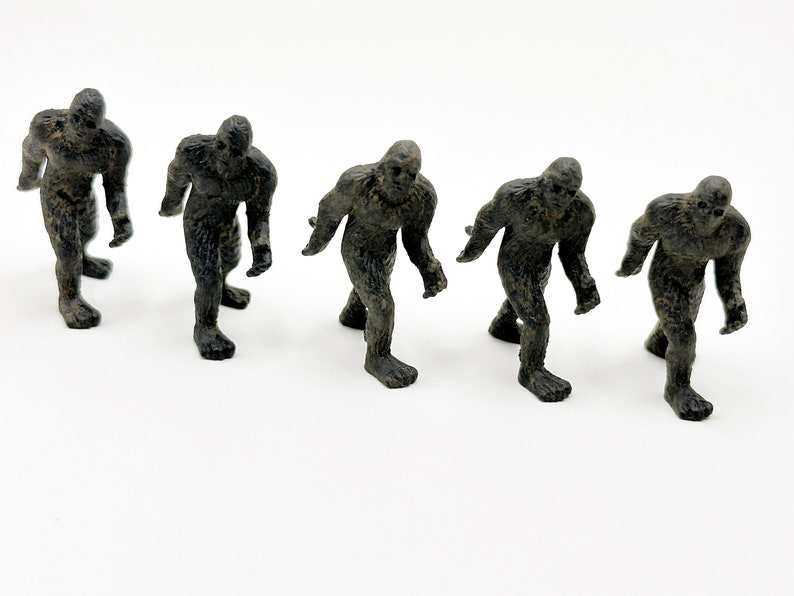 May include: Five black plastic figurines of Bigfoot, or Sasquatch, standing in a row. The figures are all facing the same direction and are in a walking pose.
