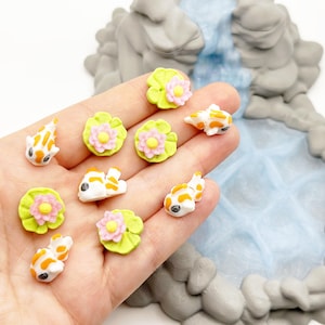 Puede incluir: Figuras de peces koi en miniatura en blanco, naranja y negro, junto con hojas de nen&uacute;far verdes con flores rosas y amarillas. Las figuras se sostienen en una mano, con una cascada en miniatura y una formaci&oacute;n rocosa en el fondo.
