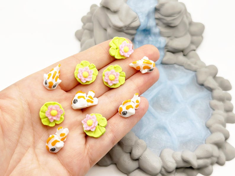 Puede incluir: Figuras de peces koi en miniatura en blanco, naranja y negro, junto con hojas de nen&uacute;far verdes con flores rosas y amarillas. Las figuras se sostienen en una mano, con una cascada en miniatura y una formaci&oacute;n rocosa en el fondo.
