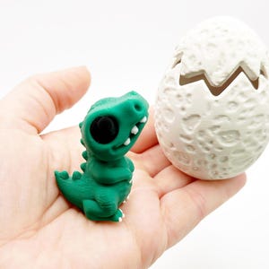 May include: A green dinosaur figurine with large black eyes and a white-toothed grin sits next to a white, cracked egg. The egg has a textured surface. The dinosaur has a spiky back and a small tail. The items are on a white background.