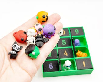 Op de afbeelding: Een verzameling kleine, kleurrijke 3D-geprinte figuurtjes waaronder een pompoen, skelet, spin en vleermuis, naast een groene plastic rasterorganizer met genummerde zwarte vierkante inzetstukken met extra kleine karakterfiguurtjes.