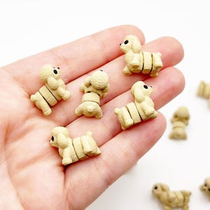 May include: A collection of small, beige, three-dimensional poodle-shaped figurines. Each miniature dog features a textured surface, rounded ears, and two distinct black dots for eyes. The figures are segmented into three parts, showing a head, torso, and hindquarters.