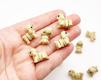 Op de afbeelding: Een verzameling kleine, beige, driedimensionale poedelvormige beeldjes. Elke miniatuurhond heeft een getextureerd oppervlak, ronde oren en twee duidelijke zwarte stippen als ogen. De figuren zijn in drie delen gesegmenteerd: kop, romp en achterwerk.