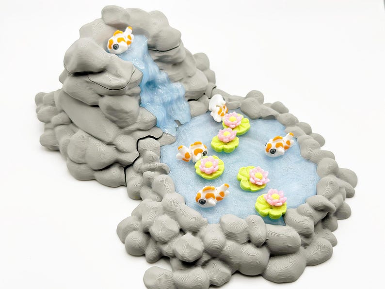 Puede incluir: Diorama en miniatura hecho a mano que representa un estanque de carpas koi con una cascada. La escena incluye formaciones rocosas grises, carpas koi naranjas y blancas, nen&uacute;fares y flores rosas. La cascada es azul claro.