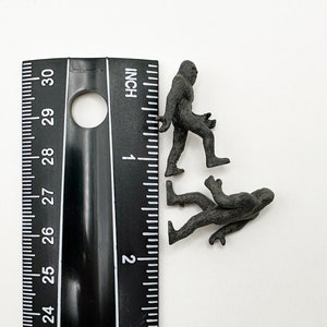 May include: Two black plastic figurines of Bigfoot, one standing upright and the other walking, next to a ruler marked in inches.