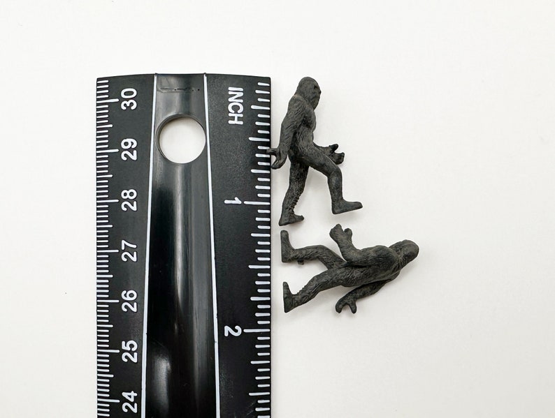 May include: Two black plastic figurines of Bigfoot, one standing upright and the other walking, next to a ruler marked in inches.