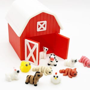 Op de afbeelding: Een rode en witte schuur met een open deur, omringd door miniatuur boerderijdieren. De dieren zijn onder meer een koe, een schaap, een varken, een kip en een paard. De schuur heeft een wit dak en deuraccenten.