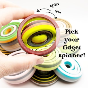 May include: A red fidget spinner with a colorful interior is held in a hand, with the word "spin" and arrows indicating movement. Other fidget spinners in various colors are in the background. Text reads "Pick your fidget spinner!"