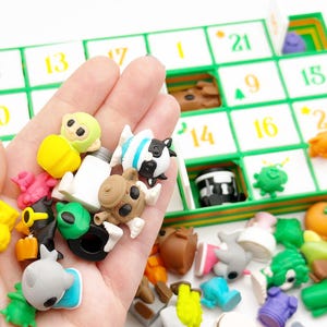 May include: A hand holding a selection of small, colourful figurines, including animal shapes and other objects. A green and white advent calendar with numbers is visible in the background. The figurines are various colours.