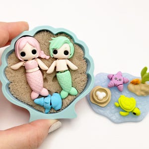 May include: A miniature ocean-themed diorama featuring two mermaid figurines with pink and green tails, set in a seashell-shaped container with sand. Additional elements include a starfish, turtle, and coral.