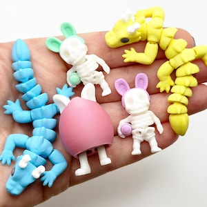 May include: Five small plastic skeleton figurines, including a blue alligator, a yellow dragon, a pink egg-shaped creature, and two bunny skeletons with pastel pink and green ears.