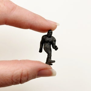 May include: A small black plastic figurine of a Bigfoot or Sasquatch walking, held between two fingers.